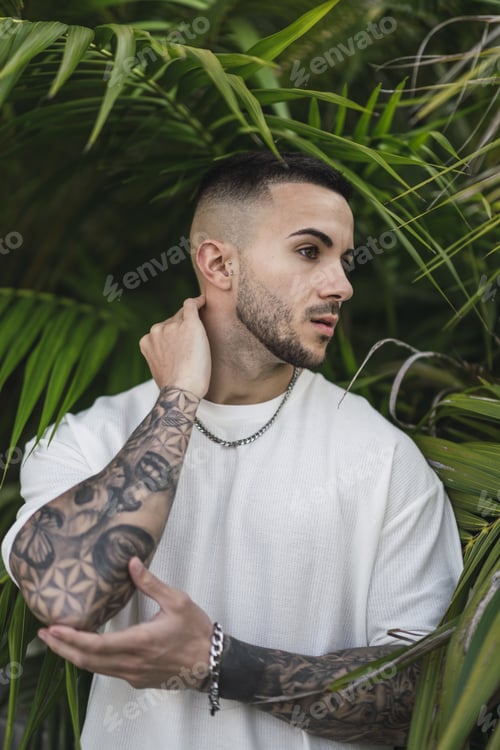 Preview: Vertical shot of a handsome Spanish Caucasian man with tattoos posing in front of green foliage