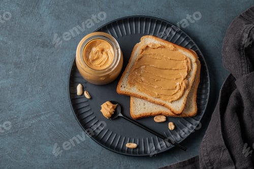Preview: Peanut butter toast on a black plate on a dark background with nuts, napkin and spoon.