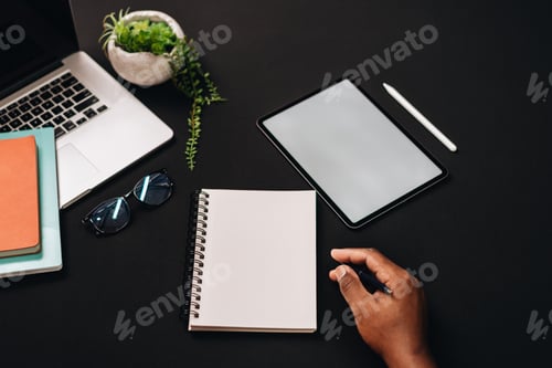 Preview: African American man sitting at black work desk with blank notebook open and tablet and laptop