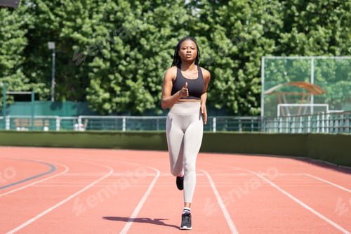 Preview: Focused sportive woman runs on red track of big stadium against city park with lush green trees