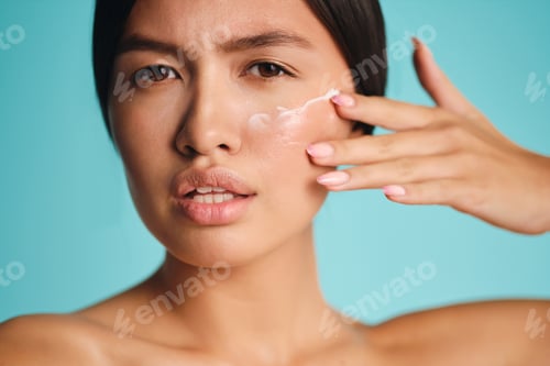 Preview: Close up attractive Asian brunette girl applying cream on clean skin intently looking in camera