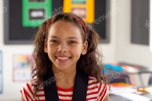 Preview: Multiracial girl standing and smiling in classroom, with red headband and bulletin board and desks