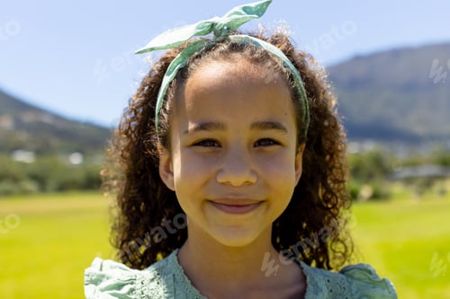Preview: Biracial girl with curly hair smiles outdoors, green headband matching her dress
