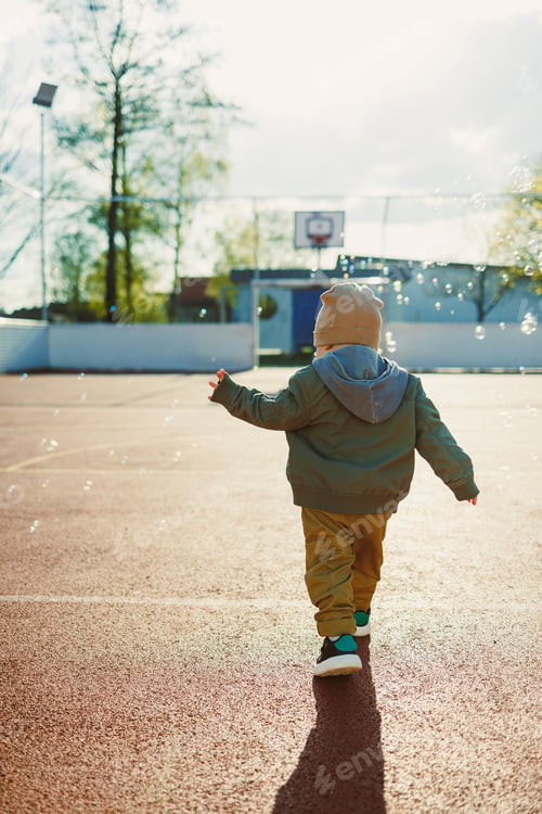 Preview: Cheerful 2 year old boy playing outside in summer in a green jacket with soap bubbles.