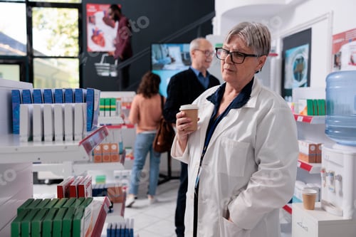 Preview: Elderly drugstore worker holding cup of coffee while looking at pharmacy shelves full with vitamins