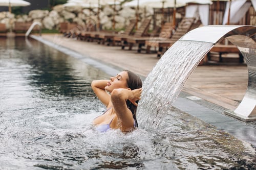 Preview: Young beautiful woman in bikini getting hydrotherapy in outdoor pool