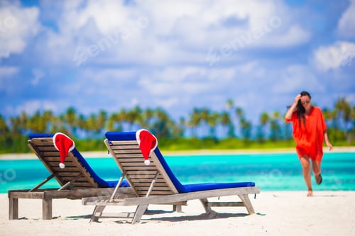 Preview: Two loungers with red Santa Hats on tropical white beach with turquoise water. Young woman walking