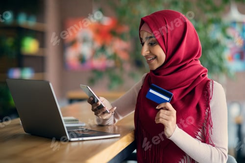 Preview: Arabic lady holding credit card, using laptop and smartphone