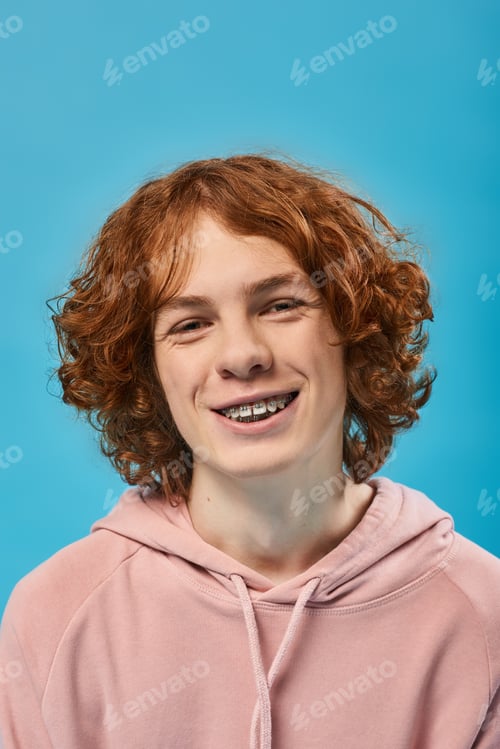 Preview: portrait of cheerful teenage guy with red wavy hair and braces smiling at camera on blue, happiness
