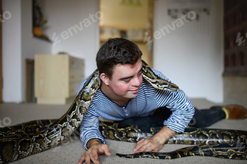 Preview: Down syndrome adult man sitting indoors in bedroom at home, playing with pet snake.