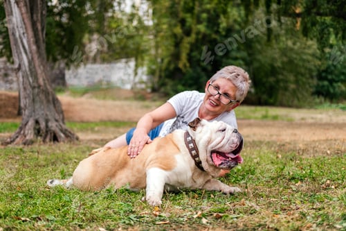Preview: Smiling senior woman laying on grass with english bulldog outdoors. Woman hugging dog in park