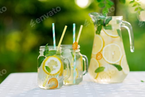 Preview: Lemonade refreshing drink in a jug and jars with lemons, fresh mint and ice on a garden table.