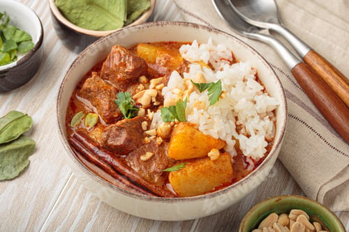 Vista previa: Cuenco de delicioso curry massaman con arroz y cacahuetes sobre fondo de madera blanca, plato tailandés