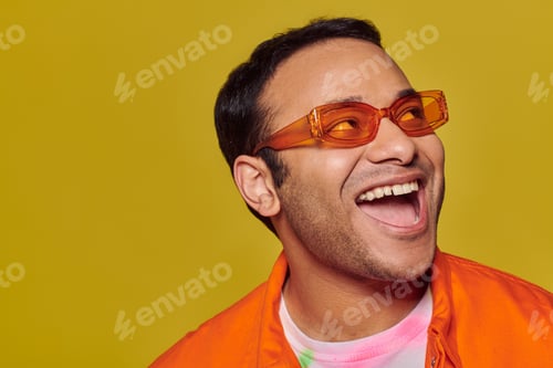 Preview: positive indian man in orange sunglasses looking away and smiling on yellow background, side glance
