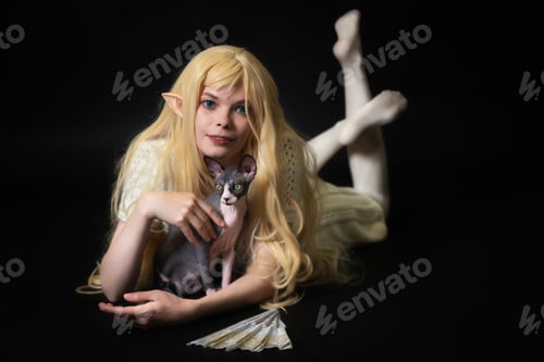 Preview: Smiling young woman elf cosplay lies on black background, caressing black and white Sphinx kitten