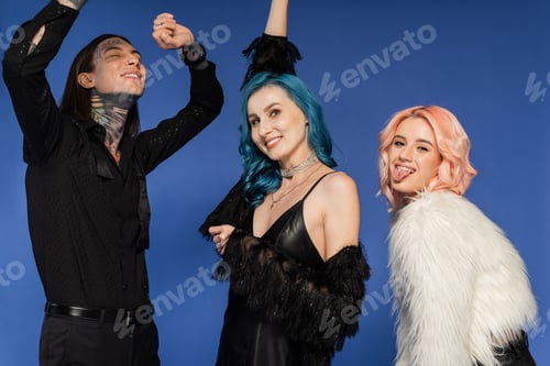 Preview: cheerful queer person sticking out pierced tongue near dancing nonbinary friends isolated on blue