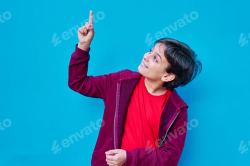 Preview: Portrait of boy in t-shirt and red jacket pointing with hand up, on blue background. Copy space