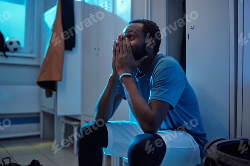 Preview: Tense young football player in uniform sitting by lockers