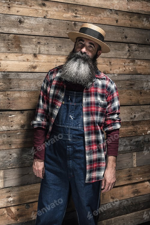 Preview: Vintage Worker Man With Long Gray Beard In Jeans Dungarees. Standing In Front Of Wooden Wall.