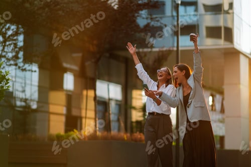 Preview: Two businesswomen celebrating success outdoors holding coffee and smartphone