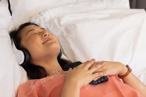 Preview: Relaxing on bed, Asian woman listening to music with headphones and holding smartphone