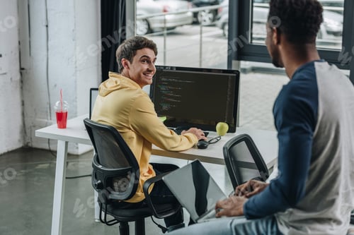 Preview: back view of african american porgrammer sitting on desk near smiling colleague