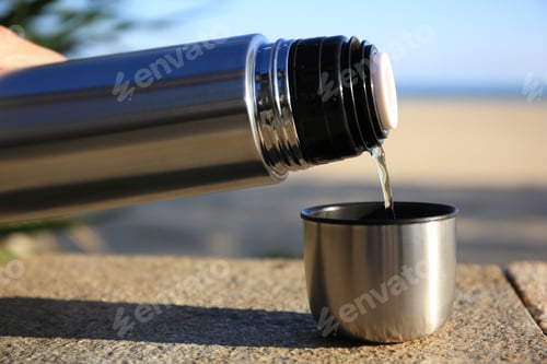 Preview: Woman pouring hot drink from metallic thermos into cap on stone surface outdoors, closeup