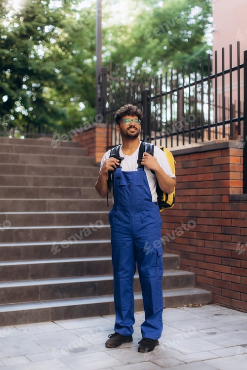Preview: Delivery man wearing blue overalls and carrying yellow backpack is standing outdoor