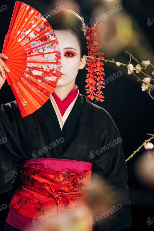 Preview: selective focus of beautiful geisha in black kimono with red flowers in hair holding hand fan in