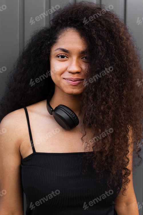 Preview: Lovely African American female with long curly hair and headphones on neck standing in city and