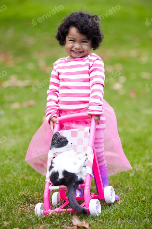 Preview: Theyre the best of playmates. Cute little girl pushing her kitten in a toy pram while outside.