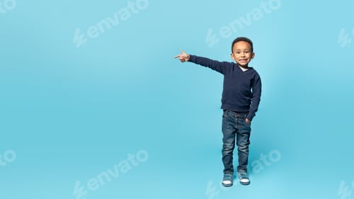 Preview: Cute african american boy looking at camera and smiling, pointing aside at free space, blue