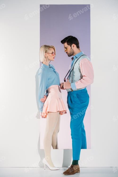 Preview: fashionable young couple in eyeglasses looking at each other while standing in aperture on grey