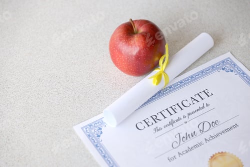 Preview: A certificate of achievement lies on table with small scroll and red apple. Education documents