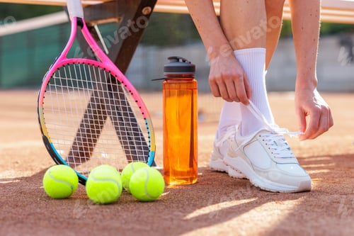 Preview: Female tennis athlete tying shoelaces on clay court with racquet balls and water bottle