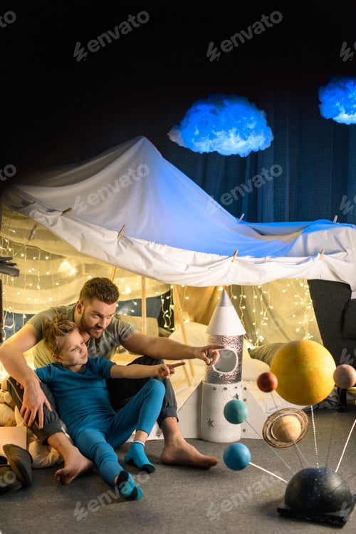 Preview: Happy father and son sitting in blanket fort together and looking away