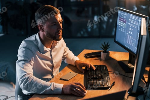Preview: Night time. Young businessman in formal clothes is in office with multiple screens
