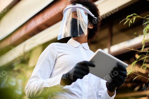 Preview: Below view of African America waitress using digital tablet while wearing face mask and visor.