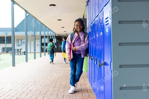 Preview: Biracial girl with a bright smile stands by blue school lockers, with copy space