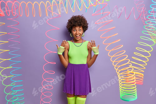 Preview: Joyful millennial girl raises hands, wears sport gloves, fashionable colorful outfit, fools around,