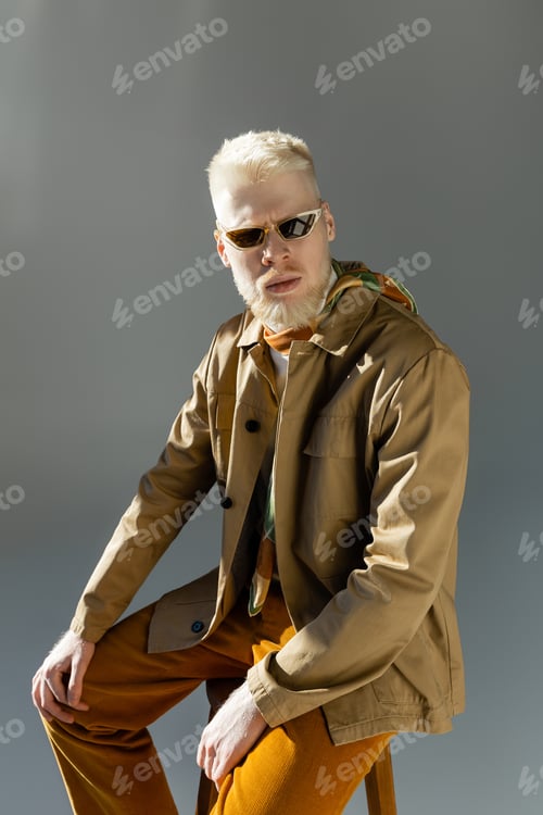 Preview: bearded albino man in stylish sunglasses and shirt jacket sitting on high chair on grey