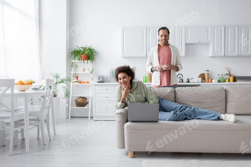Preview: Positive african american couple with laptop and coffee looking at camera in kitchen