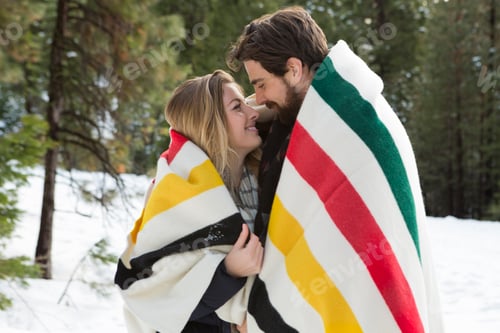 Preview: A happy couple sharing a cozy moment wrapped in a blanket against a snowy backdrop