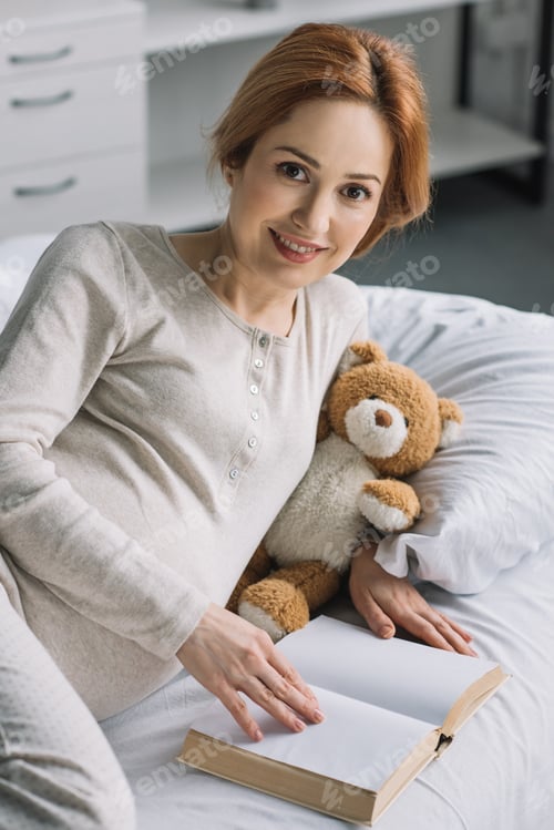 Preview: beautiful pregnant woman lying on bed with teddy bear and book