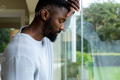 Preview: Thoughtful african american man leaning against window at home