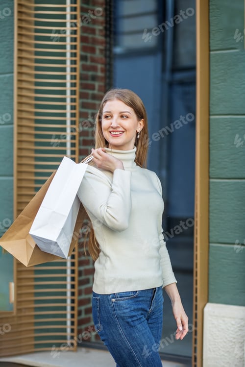 Preview: Young attractive woman holds lot of shopping bags and walks along European street.