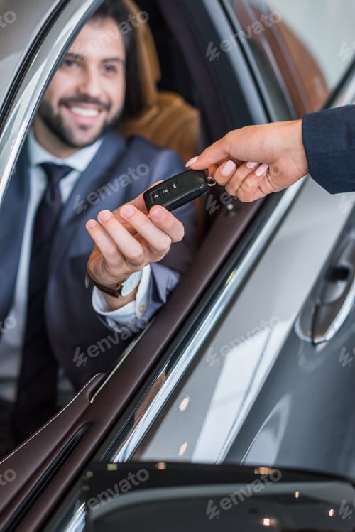 Preview: partial view of female seller giving car key to smiling businessman in new car at dealership salon