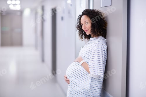 Preview: Pregnant woman leaning in hospital corridor wearing gown and wristband, cradling belly, copy space