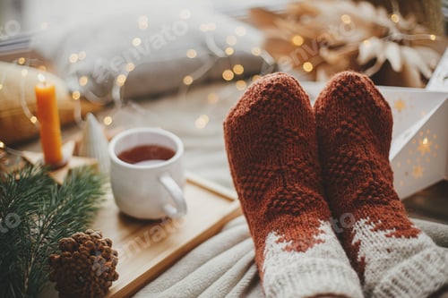 Preview: Woman feet in cozy socks, warm cup of tea, christmas star, golden lights, tree, candle. Winter hygge