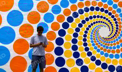 Preview: Gen Z male using technology standing on a street with a colorful art wall design in background
